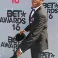 A man in a tuxedo with a purple bow tie and pocket square smiles while raising one leg and tipping his hat at the BET Awards 2016 event—CEO/CTO of DUKEWorldwide Inc., Marcus C. Duke, standing in front of a step-and-repeat backdrop.