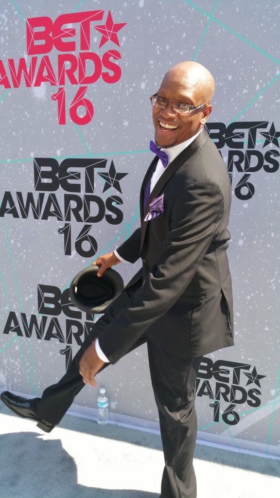 A man in a tuxedo with a purple bow tie and pocket square smiles while raising one leg and tipping his hat at the BET Awards 2016 event—CEO/CTO of DUKEWorldwide Inc., Marcus C. Duke, standing in front of a step-and-repeat backdrop.