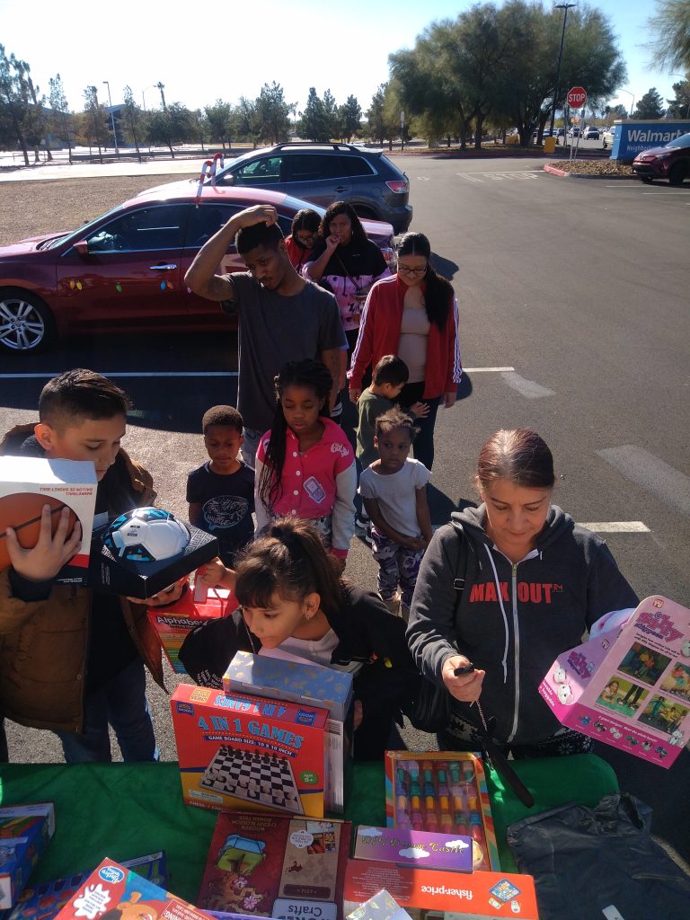 A group of adults and children gather outside in a parking lot, looking at and picking toys from a table covered with boxed games and gifts under bright sunlight at a DUKEWorldwide Inc. community event.