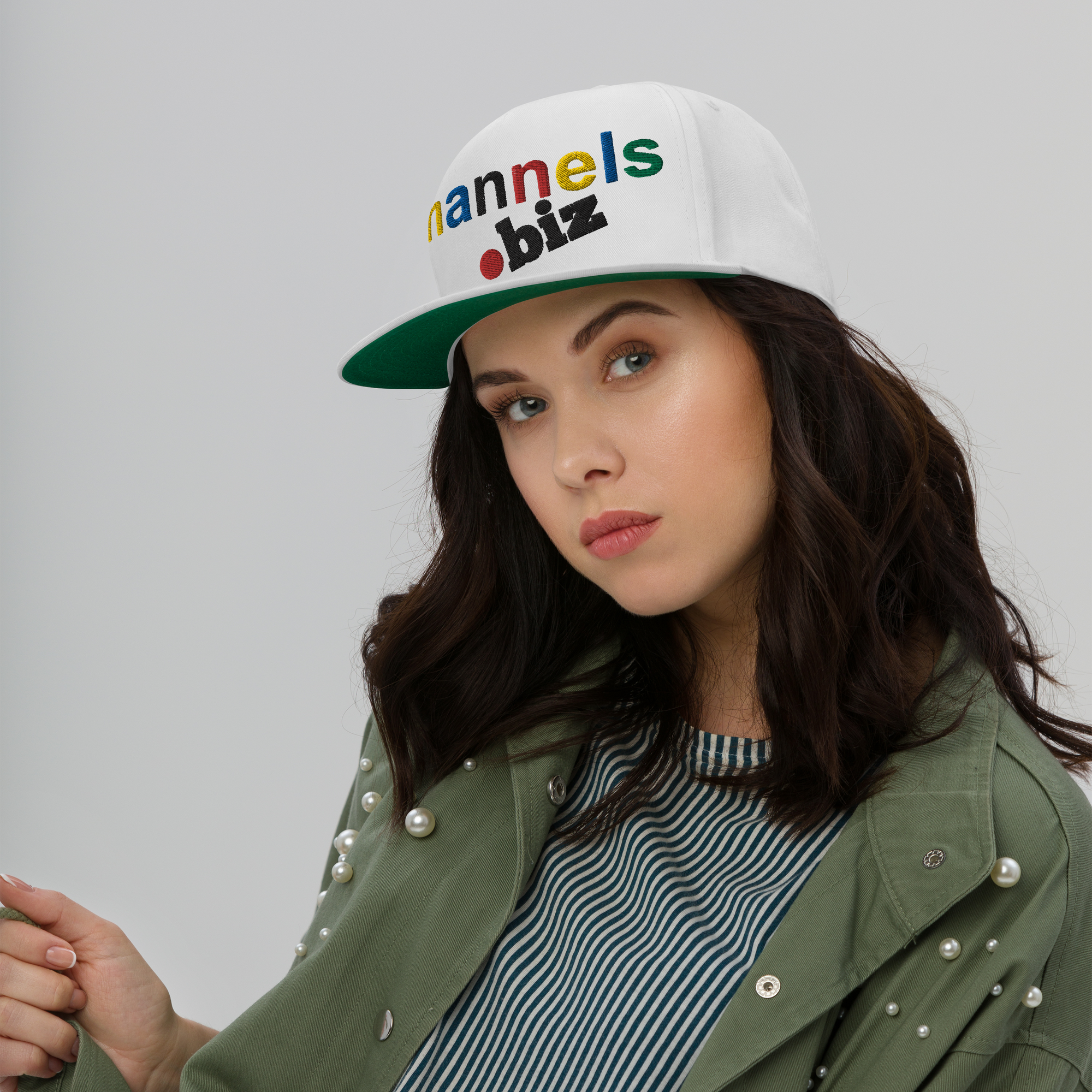 A woman with wavy brown hair wears a white cap that reads annels.biz in colorful letters. She has light makeup, a green jacket with pearls, and a striped shirt, and poses against a plain gray background.